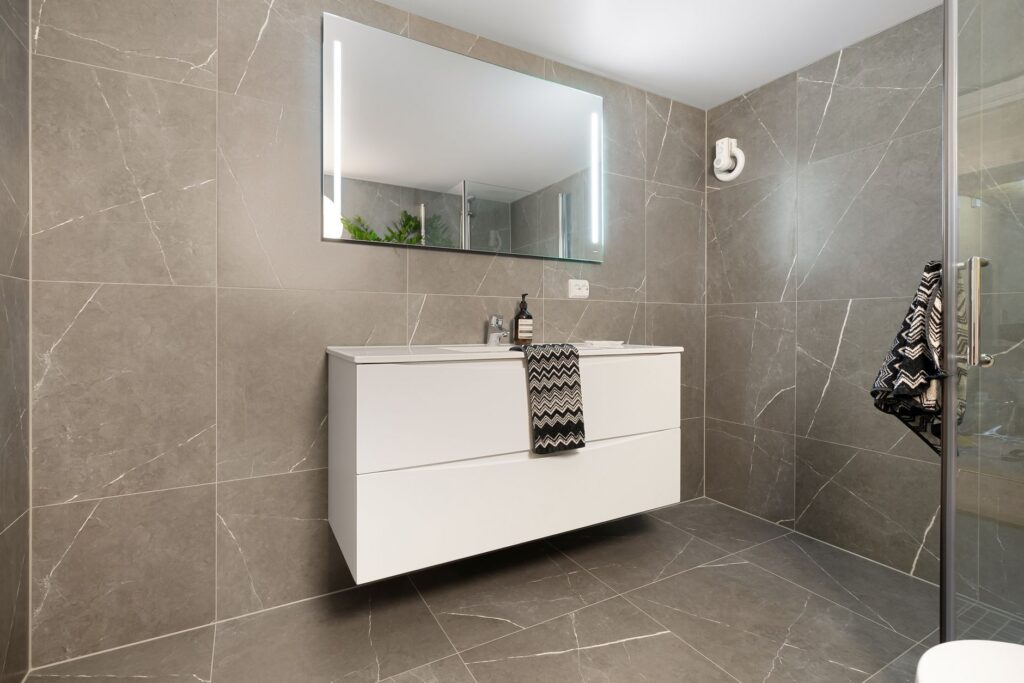 Baie modernă cu plăci ceramice mari, de culoare deschisă, și duș walk-in cu rigolă liniară