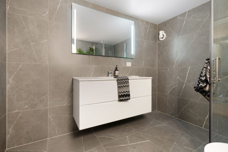 Baie modernă cu plăci ceramice mari, de culoare deschisă, și duș walk-in cu rigolă liniară